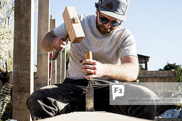 Mann mit Baseballmütze und Sonnenbrille auf der Baustelle  mit Hammer und Meißel  arbeitet an Holzbalken.