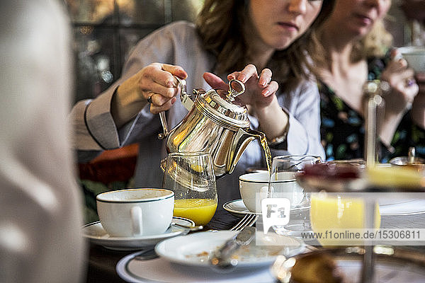 Nahaufnahme einer Frau  die an einem Tisch sitzt und Tee aus einer silbernen Teekanne gießt.