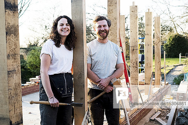 Smiling man and women holding hand tools standing on building site of residential building.