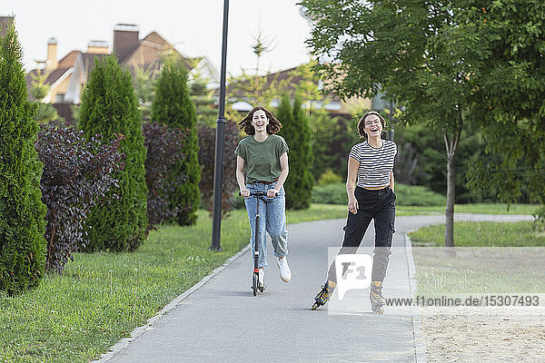 Happy women friends rollerblading and riding push scooter on sidewalk