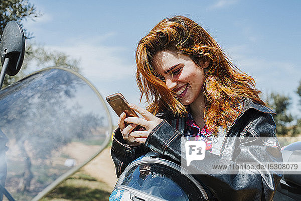 Porträt einer glücklichen rothaarigen Frau auf Motorrad mit Blick auf Handy  Andalusien  Spanien