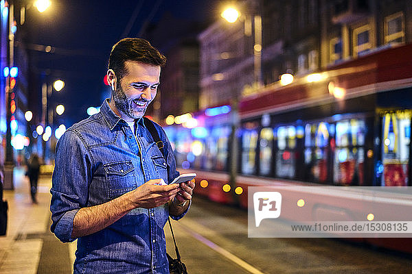 Lächelnder Mann mit kabellosen Kopfhörern mit Smartphone beim nächtlichen Warten auf die Straßenbahn
