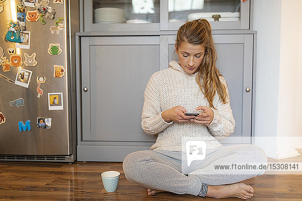 Junge Frau sitzt zu Hause in der Küche auf dem Boden und benutzt ihr Handy