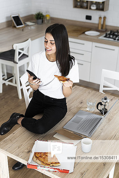Young woman with cell phone eating pizza in kitchen at home