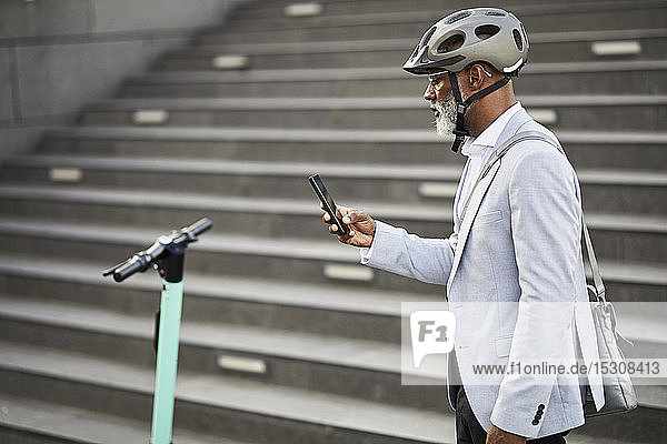 Älterer Geschäftsmann mit E-Scooter mit Fahrradhelm schaut auf Handy