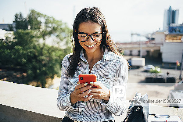 Lächelnde junge Geschäftsfrau überprüft Mobiltelefon im Freien