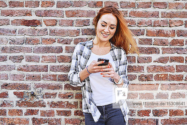 Young woman leaning against brick wall in the city while using smartphone