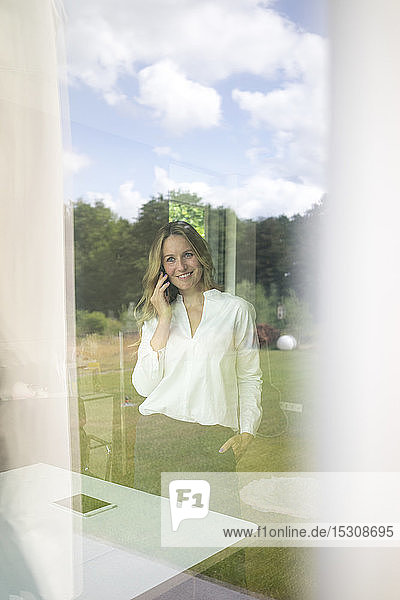 Smiling businesswoman talking on the phone at home behind windowpane
