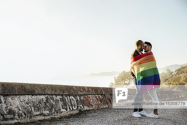 Schwules Paar  eingehüllt in eine Fahne aus schwuler Stolz  küsst sich auf einer Straße in den Bergen