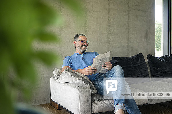 Smiling mature man sitting on couch at home reading newspaper