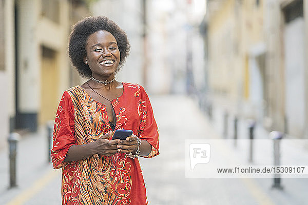 Lachende junge Frau  die mit ihrem Smartphone mitten auf der Straße steht