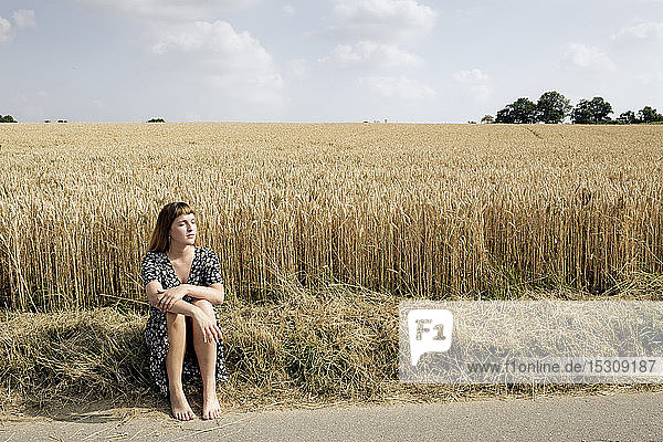 Junge Frau sitzt barfuss am Strassenrand vor einem Getreidefeld