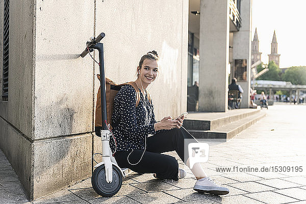Lächelnde junge Frau mit Elektroroller  Kopfhörern und Handy bei einer Pause in der Stadt