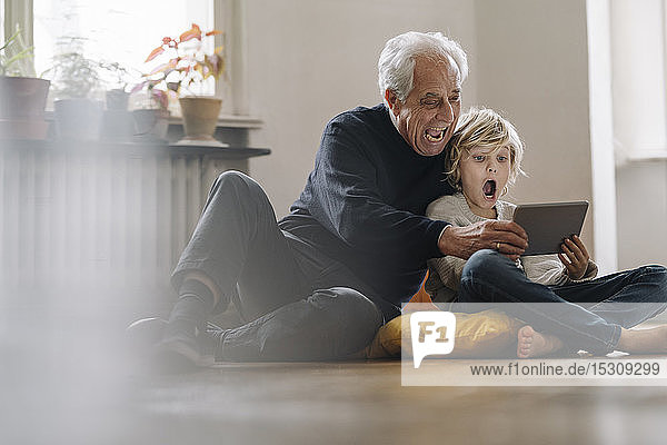 Schreiender Großvater und Enkel  die zu Hause auf dem Boden sitzen und ein Tablett benutzen