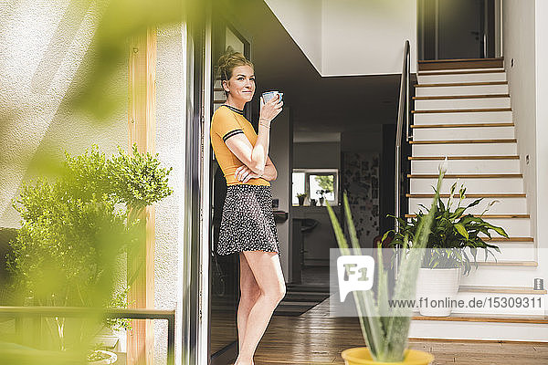 Woman holding cup of coffee leaning against terrace door at home