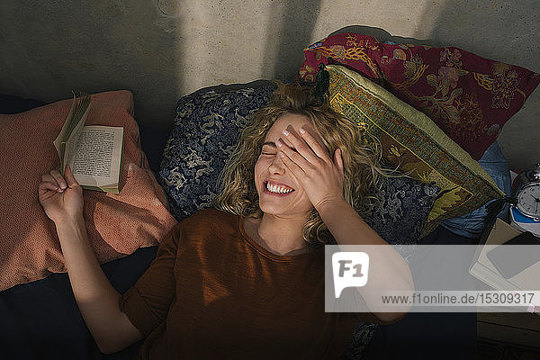 Portrait of laughing young woman lying on bed reading a book
