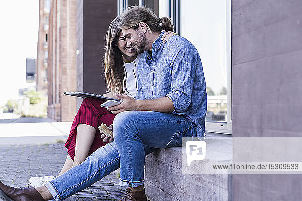 Glückliches junges Paar sitzt auf der Fensterbank eines Gebäudes und benutzt eine Tafel