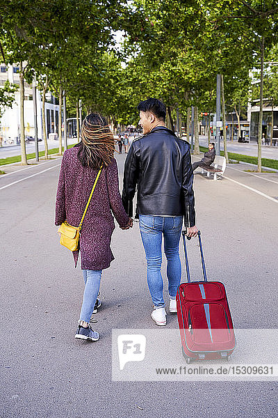 Touristenpaar  das sich an den Händen hält und mit einem Radkoffer die Straße entlang läuft  Barcelona  Spanien