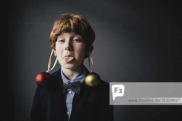 Porträt eines rothaarigen Jungen mit Fliege und Weihnachtskugeln mit herausgestreckter Zunge