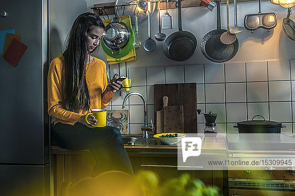Junge Frau sitzt zu Hause auf dem Küchentisch und telefoniert mit ihrem Handy