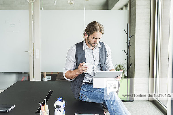 Young businessman sitting on table in office with tablet and cup of coffee