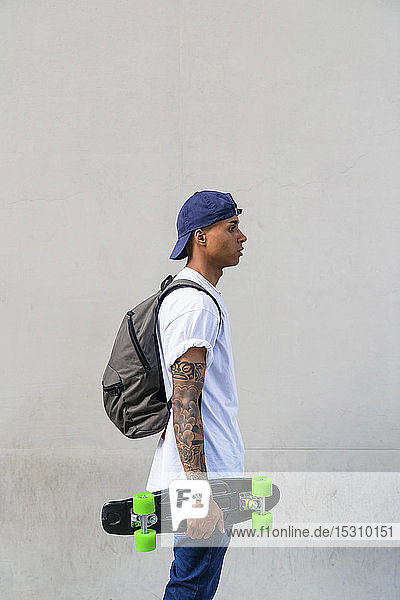 Tattooed young man with skateboard and backpack in front of grey background