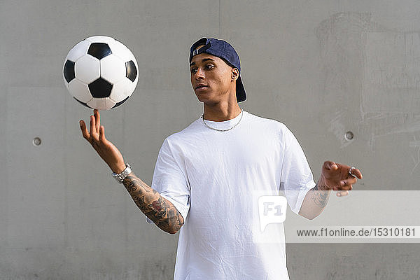 Porträt eines tätowierten jungen Mannes  der vor einer Betonwand auf seinem Finger Fußball balanciert