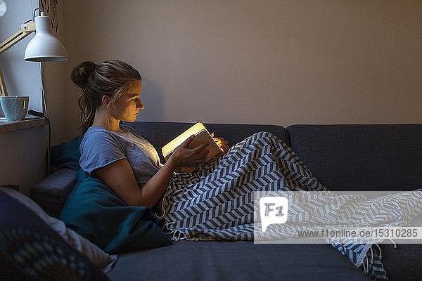 Young woman reading illuminated book on couch at home