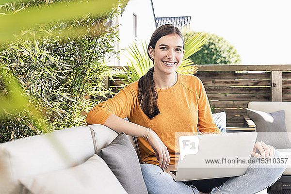 Porträt einer lächelnden jungen Frau  die mit Laptop auf der Terrasse sitzt