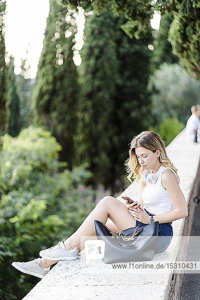 Junge Frau mit Tasche  die mit einem Smartphone an einer Wand sitzt