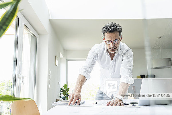 Architect working on blueprint in a bright office using laptop