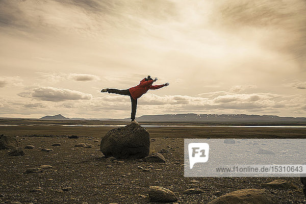 Junge Frau balanciert auf einem Bein auf einem Felsen im vulkanischen Hochland von Island