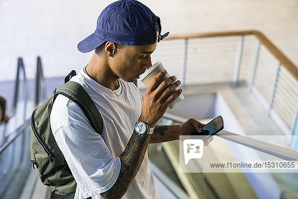 Tattooed young man with backpack standing on escalator looking at cell phone while drinking coffee to go