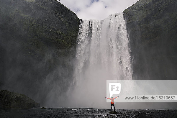 Erwachsener Mann steht mit erhobenen Armen vor den Skogafoss-Wasserfällen  Island