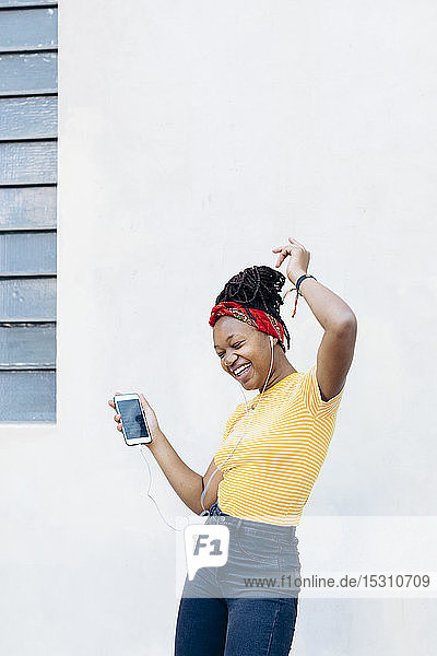 Young woman listening music with headphones and earphones singing and dancing in front of white wall