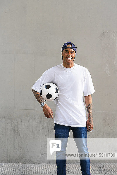 Portrait of tattooed young man with football in front of concrete wall