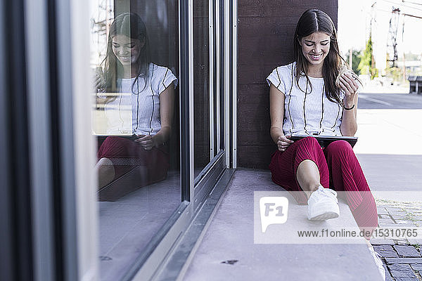Junge Frau sitzt auf der Fensterbank eines Gebäudes und benutzt ein Tablett