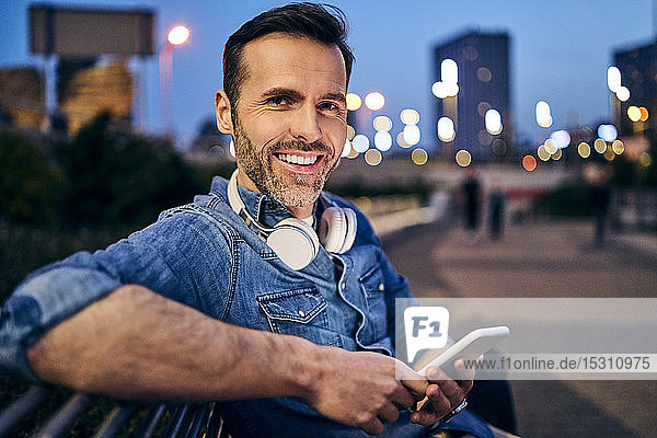 Porträt eines lächelnden Mannes  der sein Smartphone benutzt  während er abends auf einer Bank sitzt
