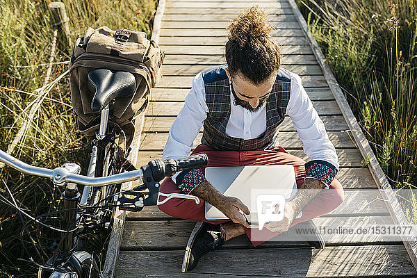 Gut gekleideter Mann sitzt auf einem Holzsteg auf dem Land neben einem Fahrrad mit Handy und Laptop