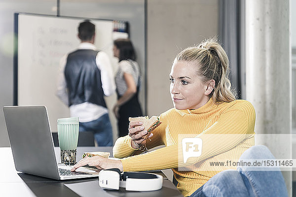 Lässige Geschäftsfrau  die im Büro mit Laptop am Tisch sitzt und ein Sandwich isst
