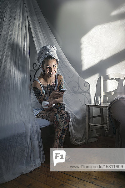 Porträt einer lächelnden tätowierten jungen Frau mit Kaffeetasse und Handy im Himmelbett