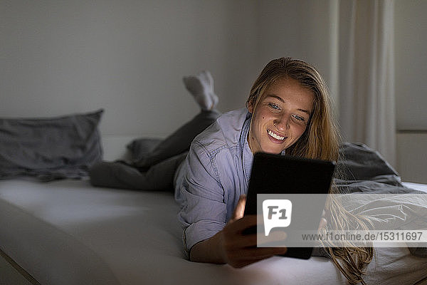 Happy young woman lying in bed at home at night using tablet