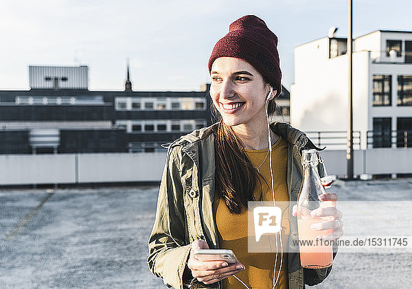 Smiling young woman with drink  earphones and cell phone on parking deck