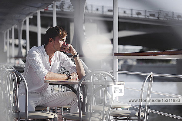 Nachdenklicher Mann in weißer Kleidung sitzt auf Ausflugsboot im Sonnenlicht und schaut in die Ferne  Moskau  Russland