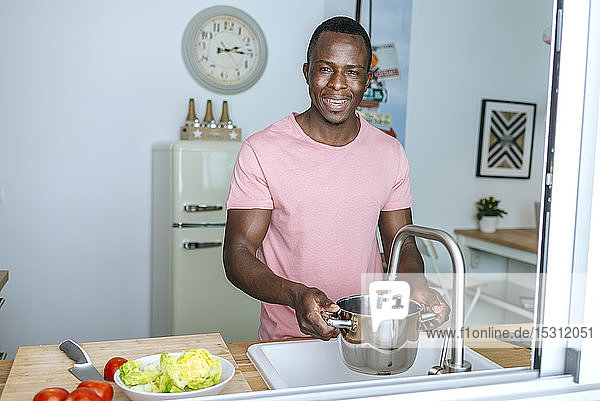 Porträt eines lächelnden jungen Mannes  der in der Küche Wasser in einen Topf gießt