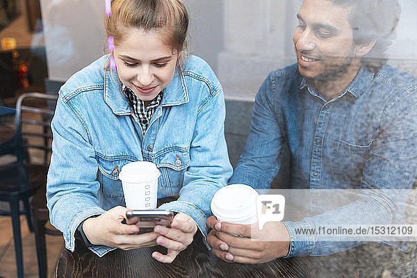 Junge Frau benutzt ein Smartphone in einem Café  während ihr Freund sie beobachtet