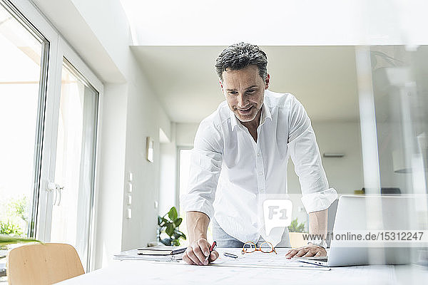 Architect working on blueprint in a bright office using laptop