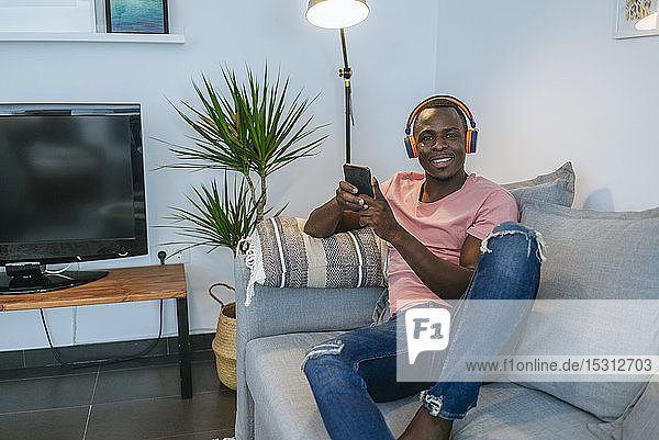Porträt eines lächelnden jungen Mannes mit Kopfhörer und Smartphone auf der Couch zu Hause