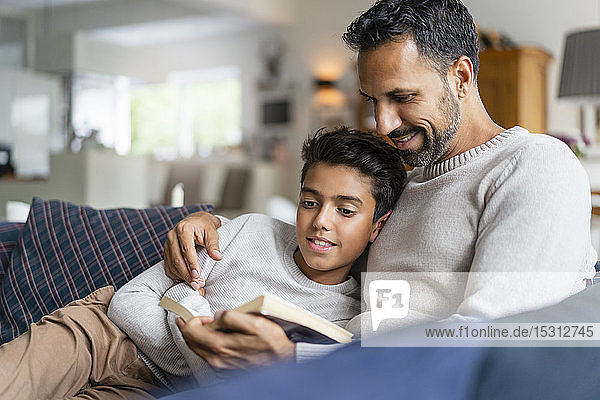 Vater liegt mit Sohn auf Couch im Wohnzimmer und liest Buch