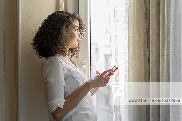 Junge Frau mit Handy  die zu Hause aus dem Fenster schaut
